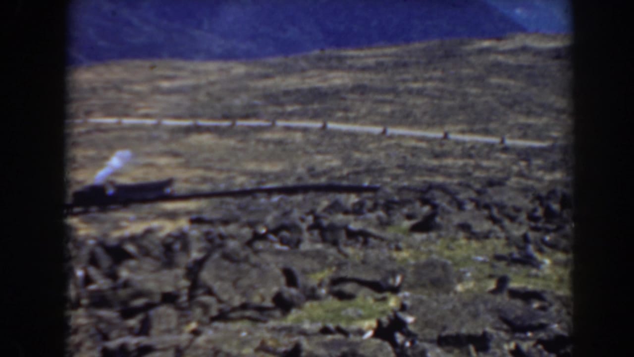 Small steam train on mountain tracks with a pale railing across the hillside.