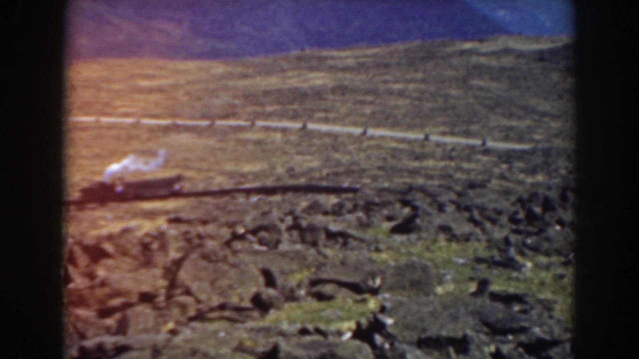 Steam train on a mountain rail line with rocks, smoke, and distant ridges.