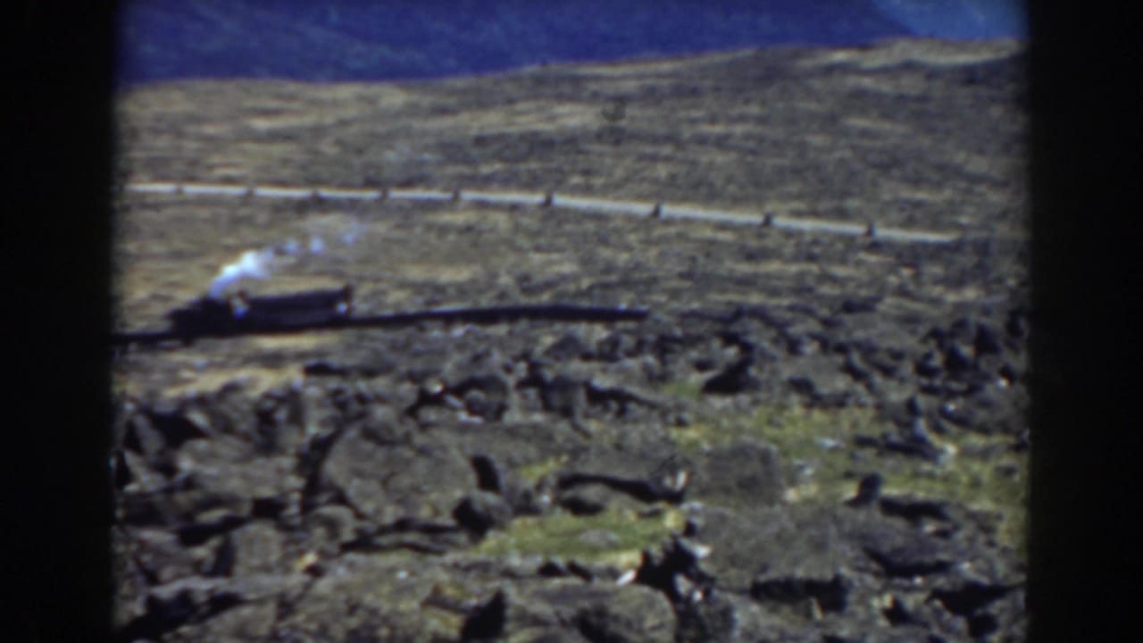 Steam train and smoke plume on tracks across a rocky hillside.