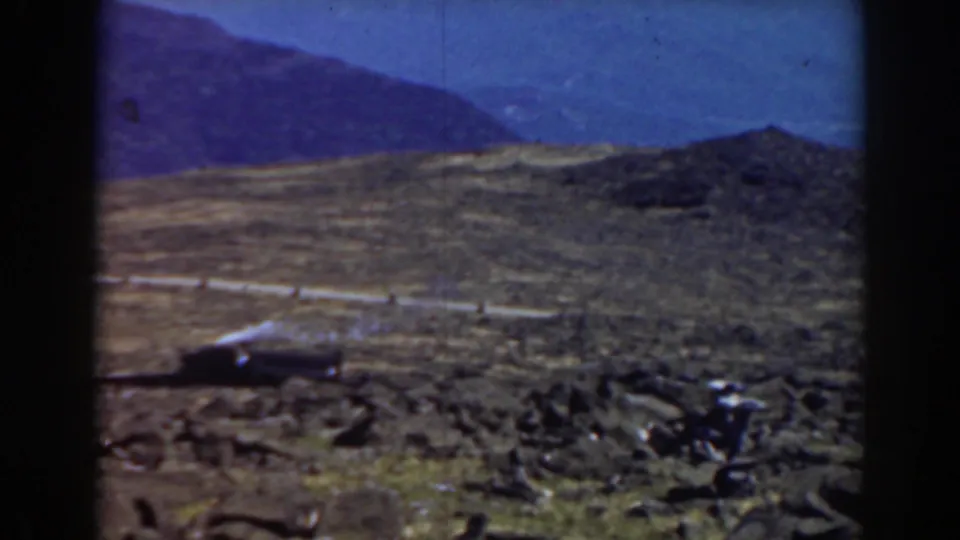 1938 Mount Washington frame showing the train farther left on the slope, with rocky terrain and mountain ridges beyond.