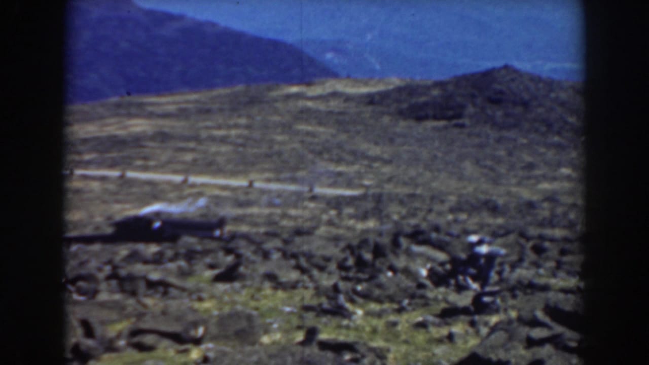 Rocky mountain terrain with a road, dark vehicle shape, distant ridges, and blue sky.