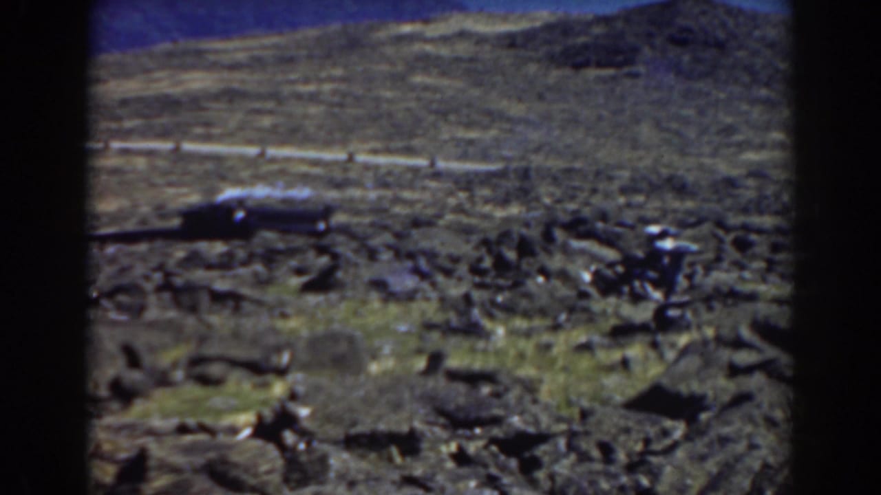 Rock-strewn hillside with a road, small dark vehicle, sparse plants, and distant mountains.