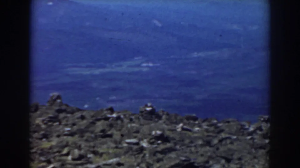 1938 Mount Washington home-movie frame showing a rocky summit edge with hazy blue valley and distant mountain landscape beyond.
