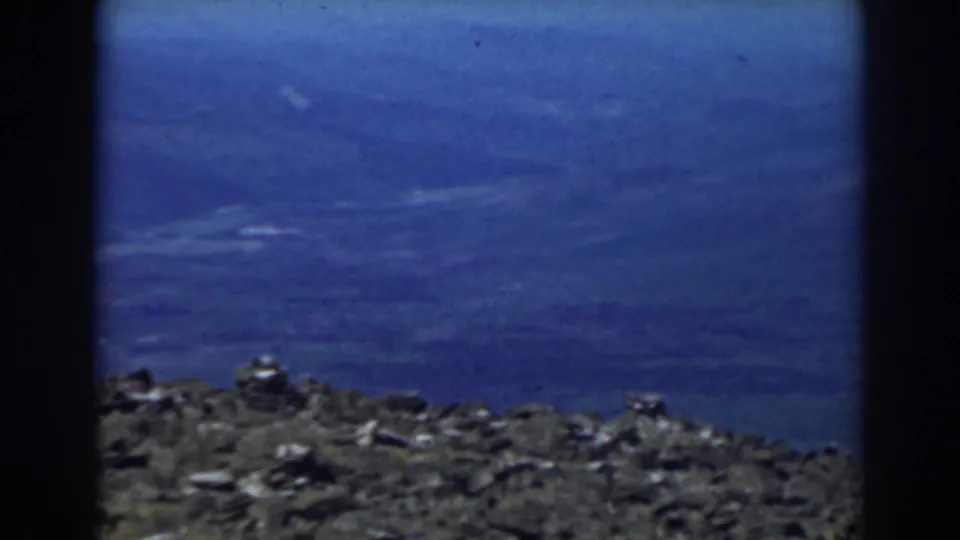 1938 Mount Washington view with rough summit rocks along the lower frame and a hazy blue sky above.