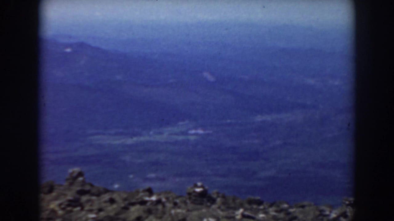 1938 Mount Washington view with a jagged rocky ledge below layered blue mountain slopes and atmospheric haze.