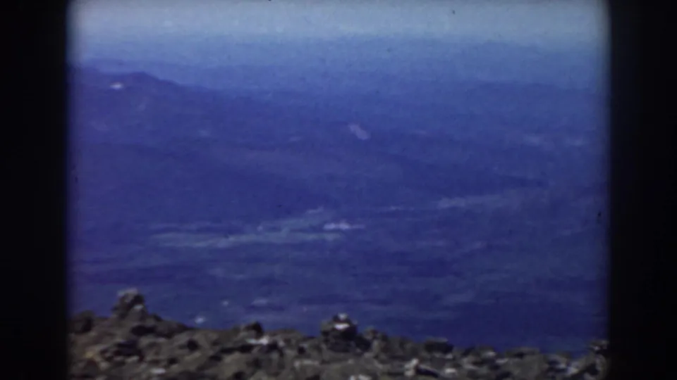1938 Mount Washington view with a jagged rocky ledge below layered blue mountain slopes and atmospheric haze.