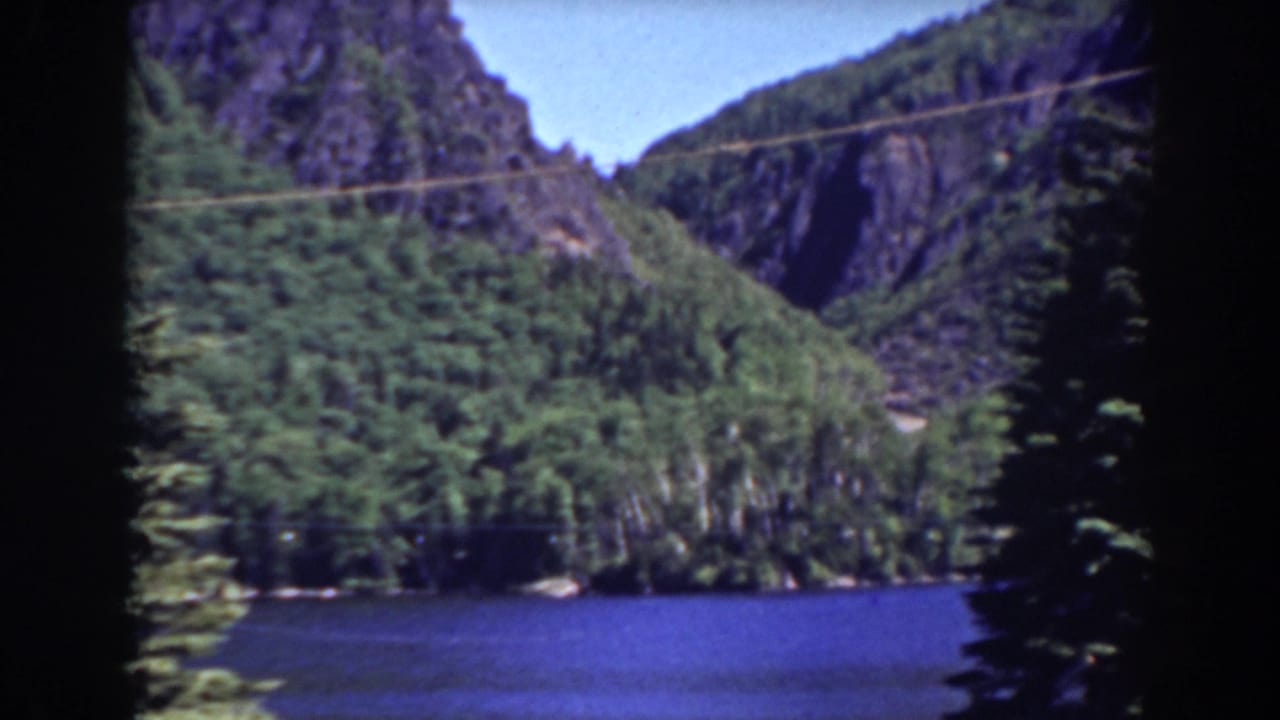 Forested mountain slopes above a blue lake with rocky cliffs and dark film edges.