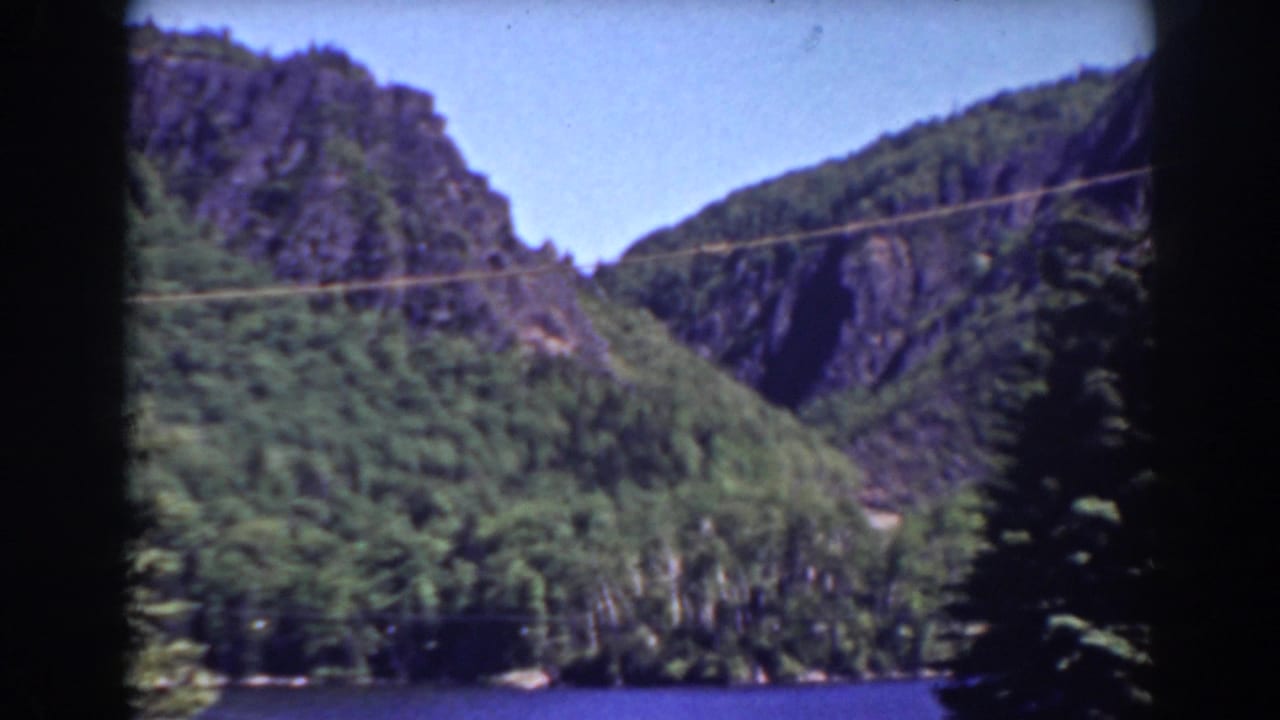 Mount Washington-area home-movie frame with a forested lakeshore, steep rock faces, and blue sky, partly obscured by dark frame edging.