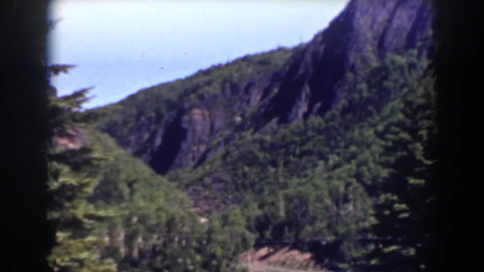 1938 8mm scenic frame from the Mount Washington area, showing dense trees below steep exposed rock and pale blue sky.