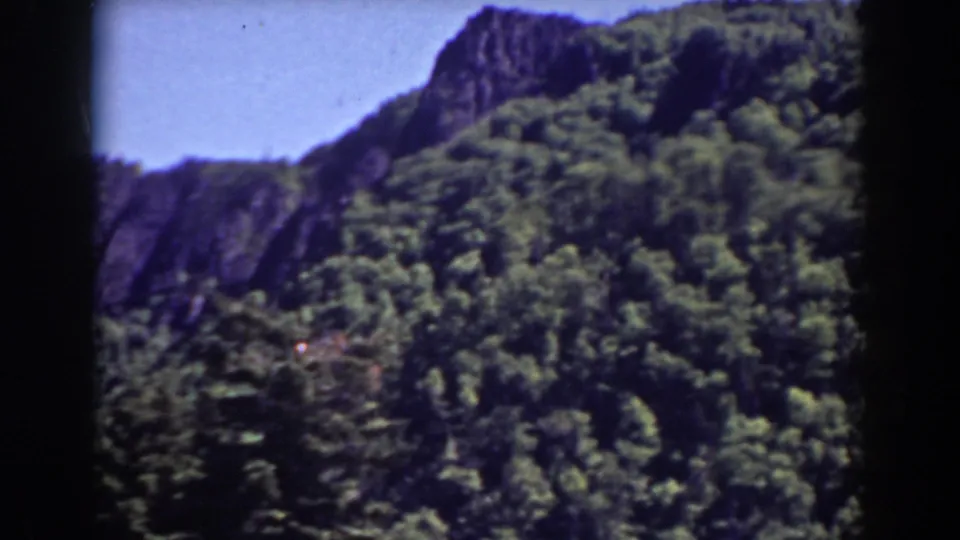 A 1938 8mm home-movie frame of Mount Washington with dense green forest, rocky outcrops, blue sky, and soft small-gauge film texture.