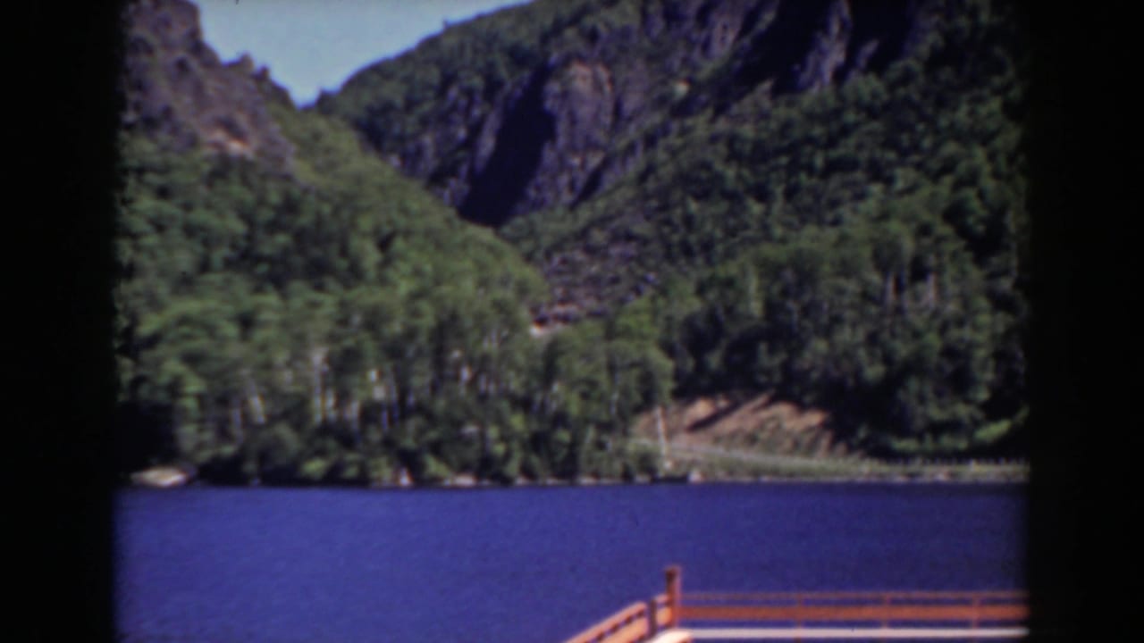 Mount Washington-area home-movie frame showing calm blue water, wooded shoreline, and steep rocky slopes beyond a dock railing.