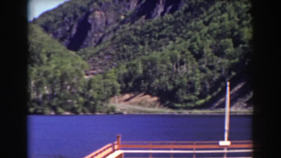 1938 home-movie view of a calm mountain waterway with shoreline trees, rocky slopes, and dock railings in the foreground.