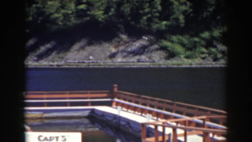 1938 8mm lakeside view with a dock corner, partial 'CAPT S' sign, blue water and forested rocky slope.