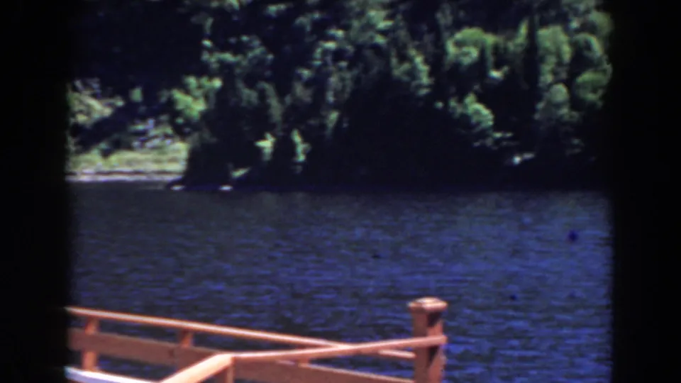1938 Mount Washington, New Hampshire frame emphasizing dock railing, calm water, and shaded trees, with the sign no longer centered.