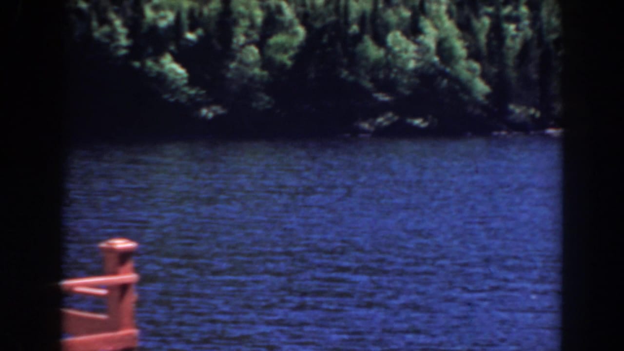 Blue lake and forested shore with a reddish railing post near the lower frame.