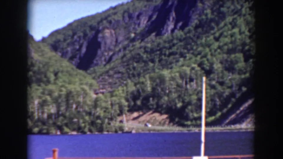 1938 8mm Mount Washington area view of a forested hillside and rocky slope above a blue lake, with railing and post in the foreground.