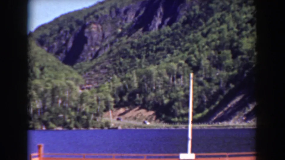 1938 New Hampshire home-movie frame showing lake water, forested slopes, rocky hillside, railing, and a white pole.