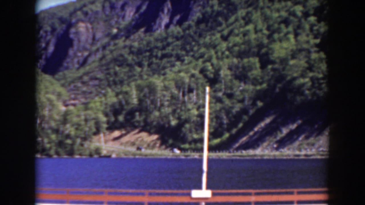 1938 New Hampshire frame with blue water, dense green hillside, exposed rock, and foreground railing from a visitor viewpoint.
