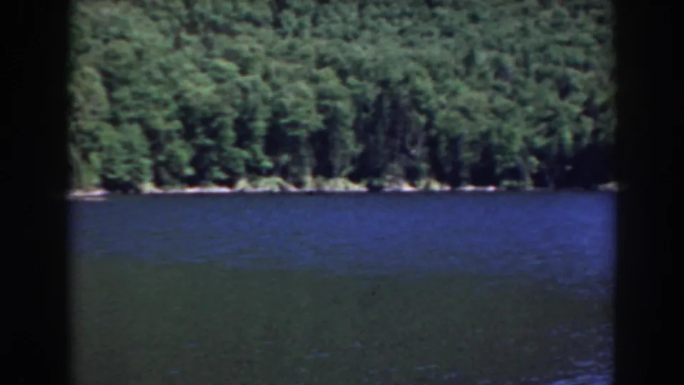 Vintage 8mm frame showing rippled lake water and a continuous green forest shoreline in the Mount Washington, New Hampshire area.