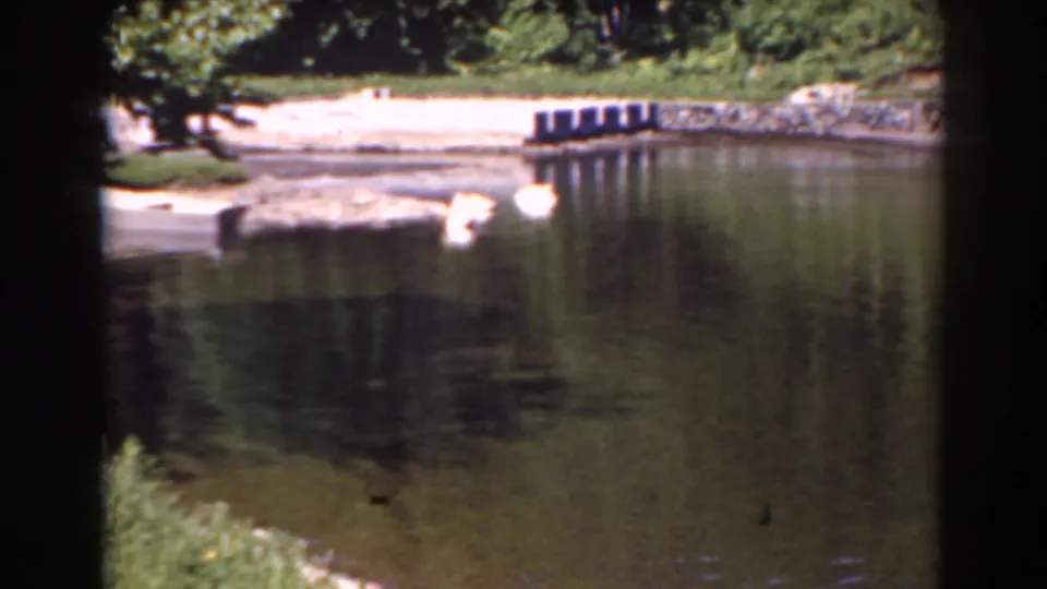 1938 home-movie frame showing a quiet lakeshore with beach, stone edging, water reflection, and dark dock-like posts.