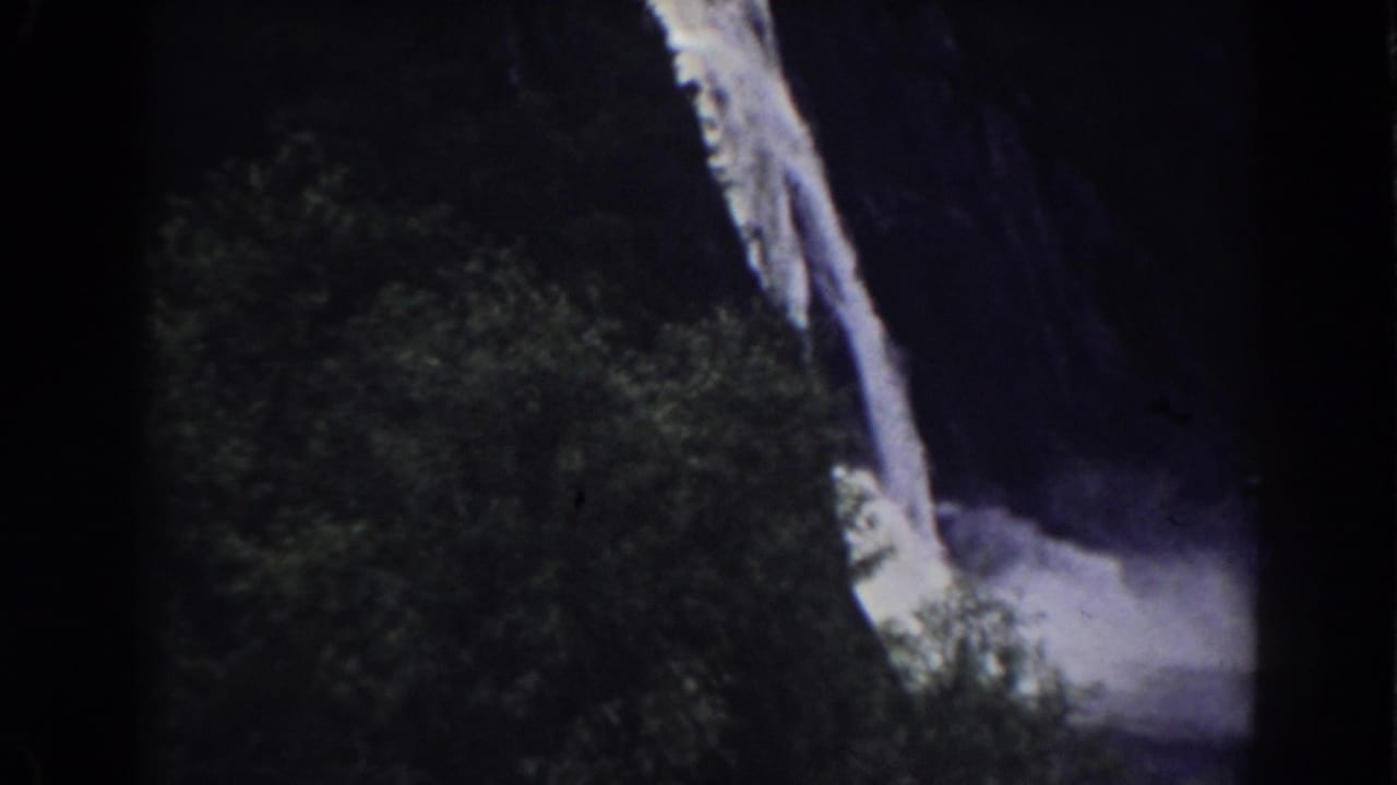 Waterfall drops beside dense trees into mist at the base of a dark rock face.