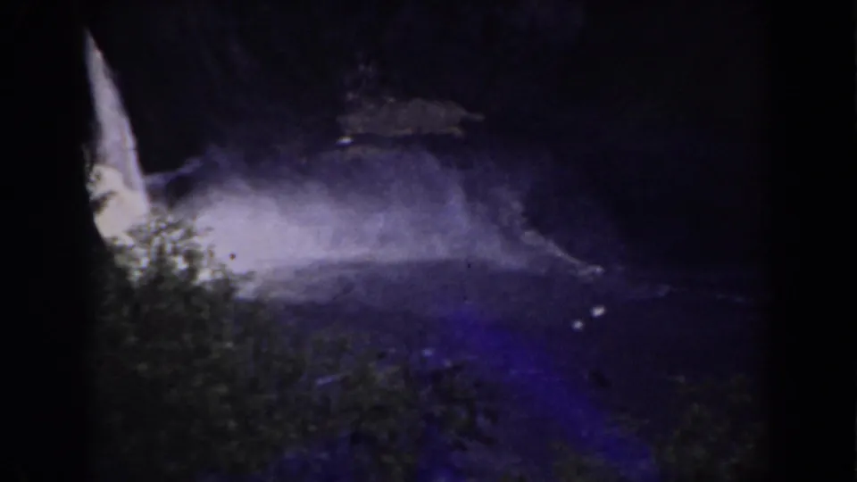 The waterfall is framed lower in the shot, with foreground foliage and pale mist crossing the dark pool.