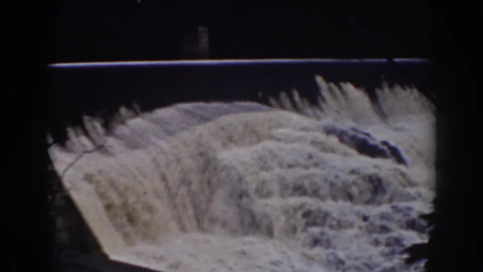 The 8mm frame shows turbulent water descending a dam spillway, with dark trees and the sloped edge of the structure visible around it.