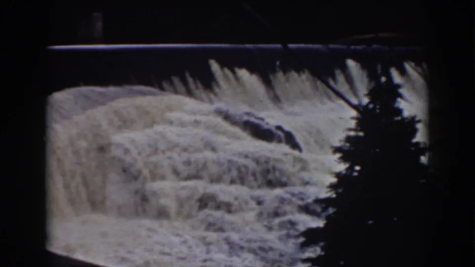 Foamy water continues over the Mount Washington dam spillway, partly obscured by dark tree silhouettes in the foreground.