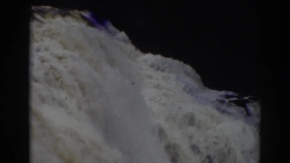 Close 8mm frame of Crystal Cascade water moving over rough rock, with faded color and heavy shadow around the film gate.