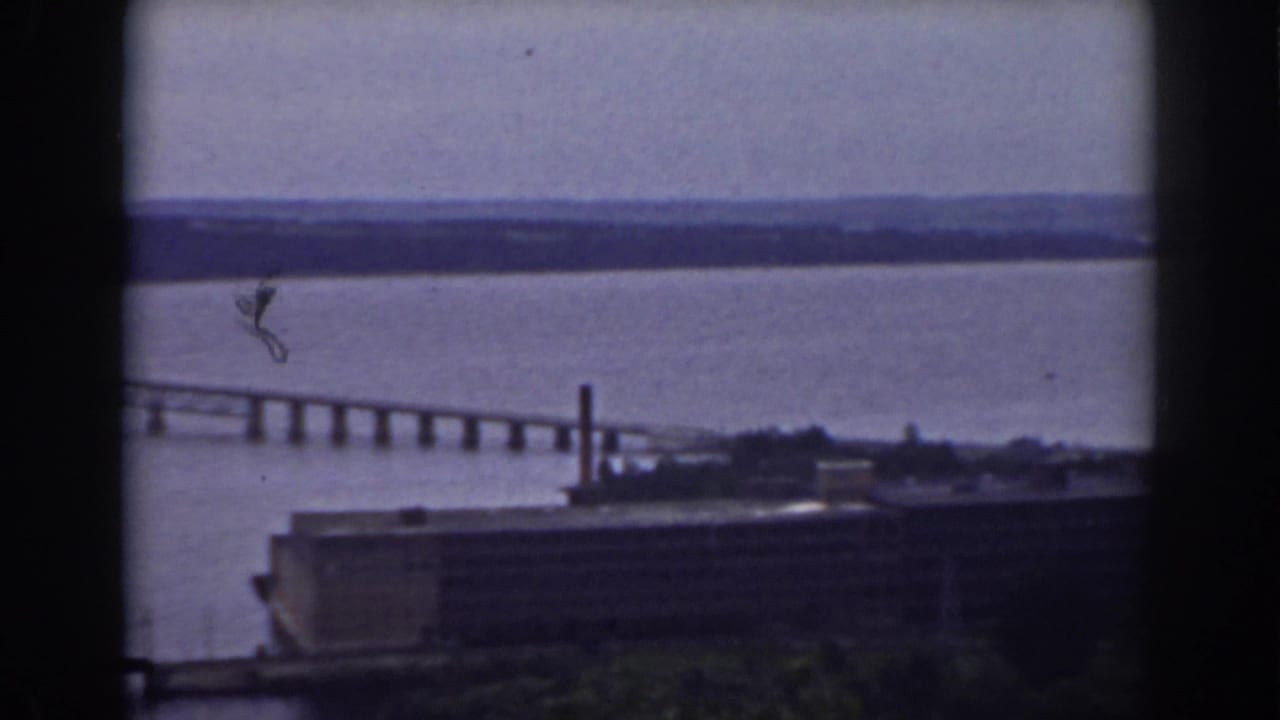 Long bridge crossing a wide river with a low industrial building in the foreground.