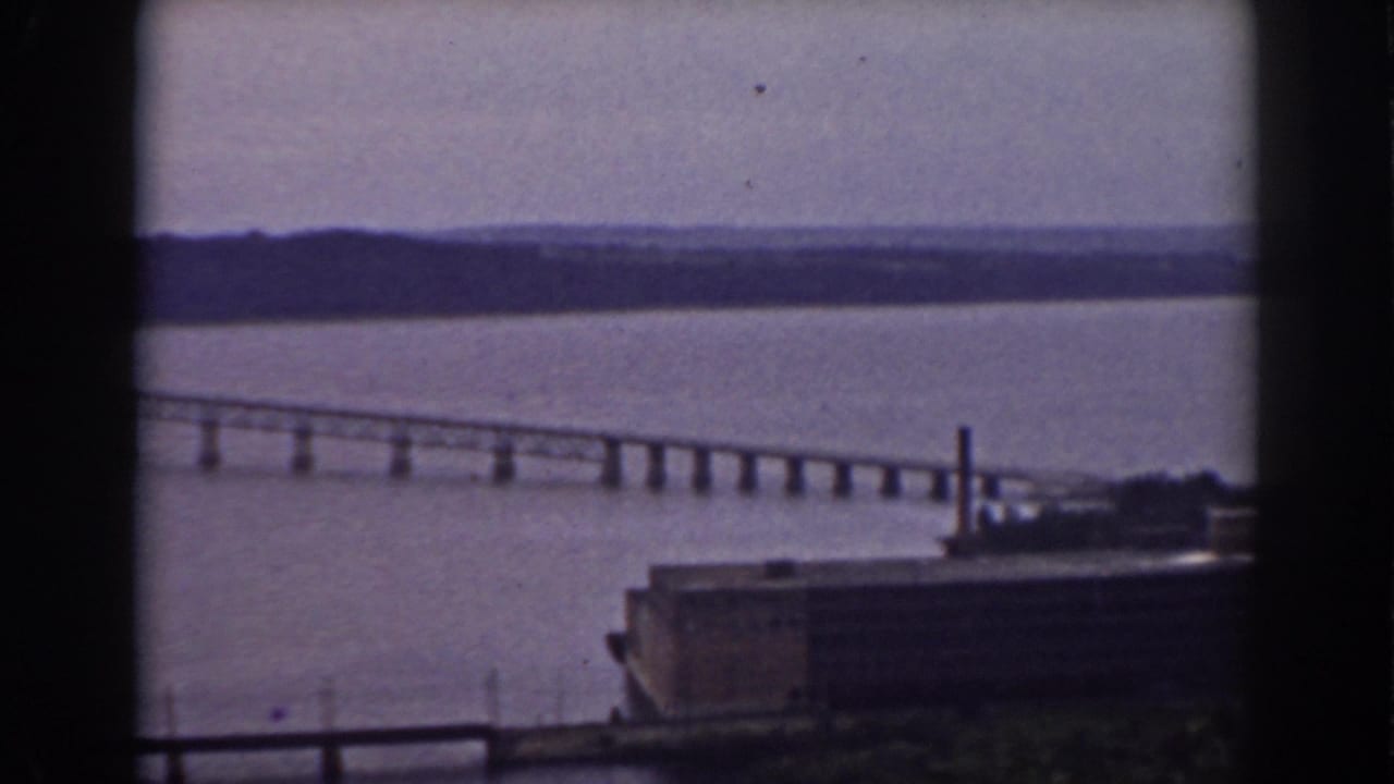 A long bridge crosses the river beyond an industrial waterfront structure, framed by dark interior window edges.