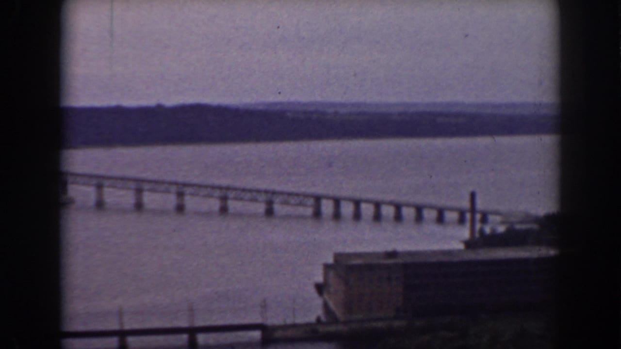 Late-1930s home-movie frame showing a long river bridge, distant shoreline, and industrial foreground near Mount Washington, New Hampshire.