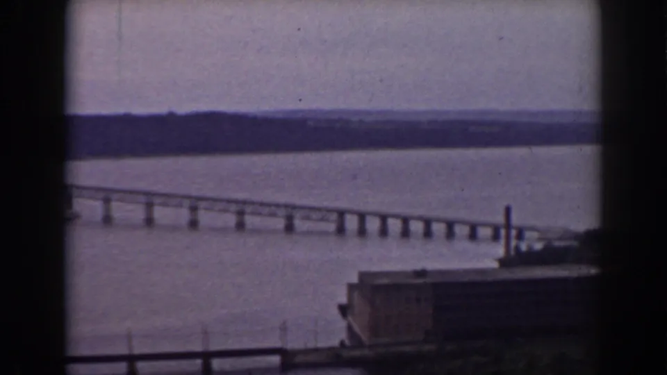 Late-1930s home-movie frame showing a long river bridge, distant shoreline, and industrial foreground near Mount Washington, New Hampshire.