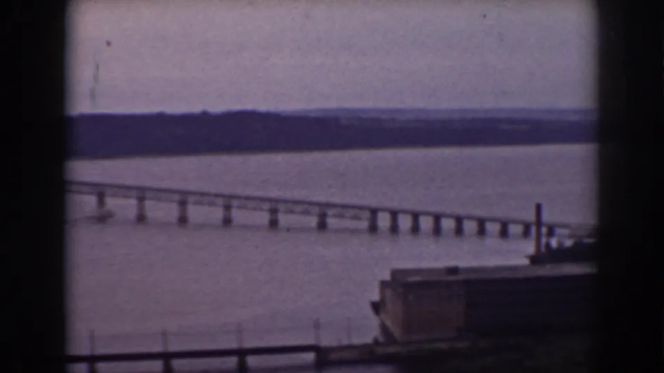 1938 New Hampshire home-movie frame showing a long bridge over a lake, distant shoreline, and dark window bars in the foreground.