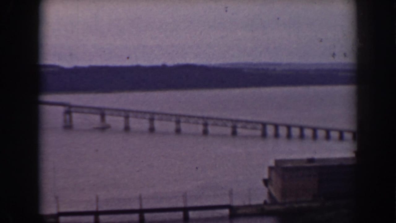 Window-framed 1938 New Hampshire view of a bridge stretching across a broad lake beneath a muted sky.