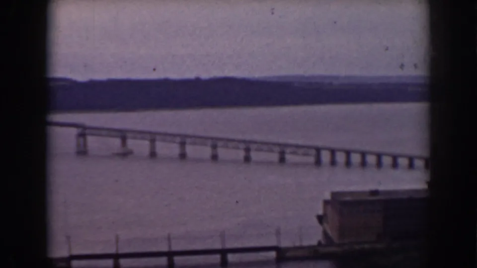 Window-framed 1938 New Hampshire view of a bridge stretching across a broad lake beneath a muted sky.