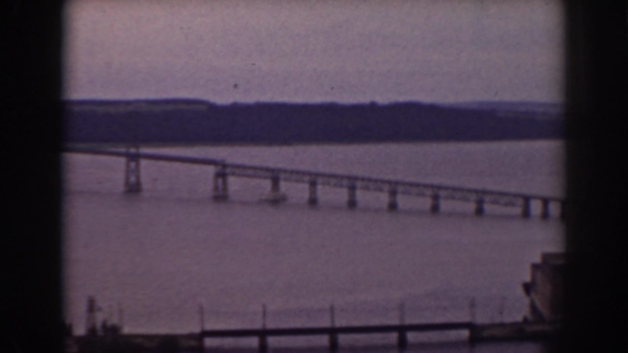 1938 home-movie still showing a lake bridge, dark foreground window bars, distant tree line, and partial structure at the right edge.