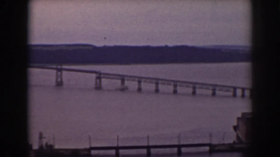 1938 home-movie still showing a lake bridge, dark foreground window bars, distant tree line, and partial structure at the right edge.
