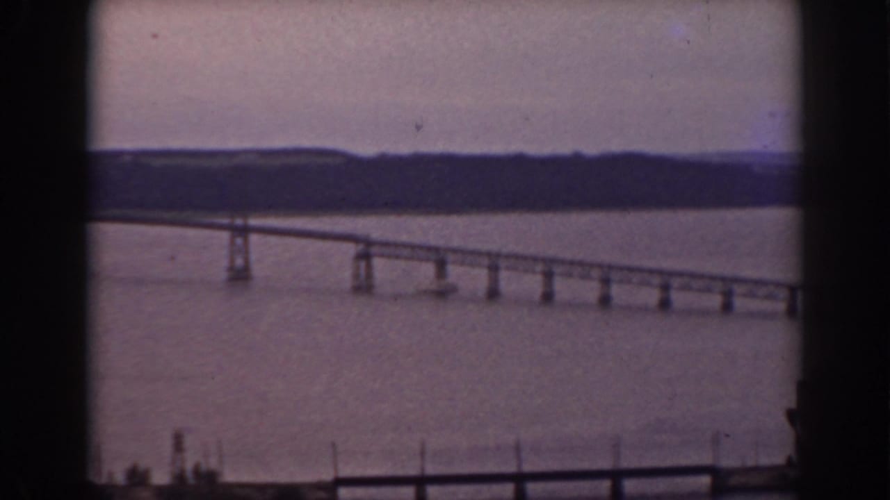 Long bridge crossing broad water, seen through a dark window frame with a wooded shore beyond.