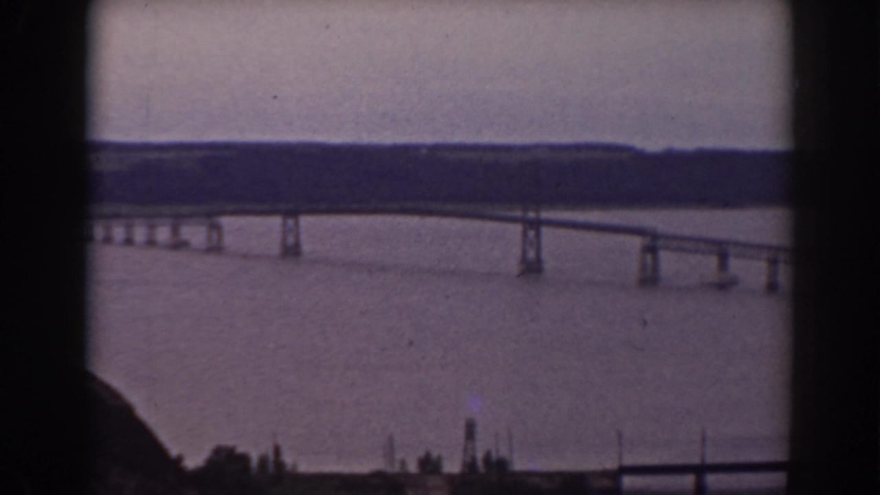 The bridge recedes across the water in a 1938 home-movie view, with dark window framing and a muted wooded shoreline in the distance.