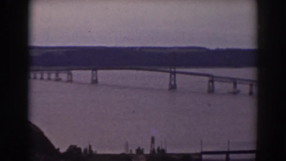 The bridge recedes across the water in a 1938 home-movie view, with dark window framing and a muted wooded shoreline in the distance.