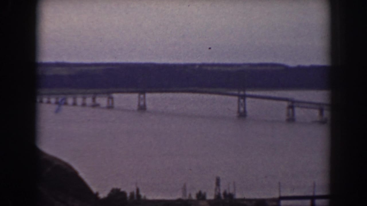 Long bridge crossing wide water, seen from a dark rocky overlook beneath an overcast sky.