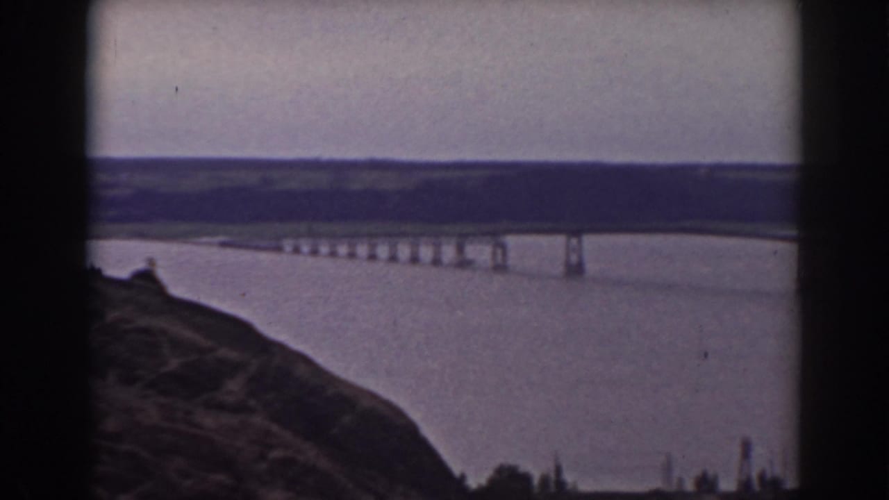 Final 1938 New Hampshire frame of the bridge and water landscape, viewed from a high rocky foreground with distant hills.
