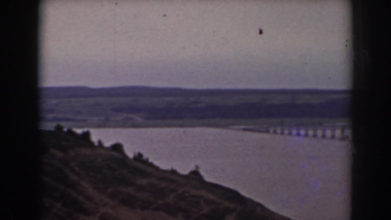 Elevated lake view with bridge at right, sloped foreground, distant hills, and muted sky.