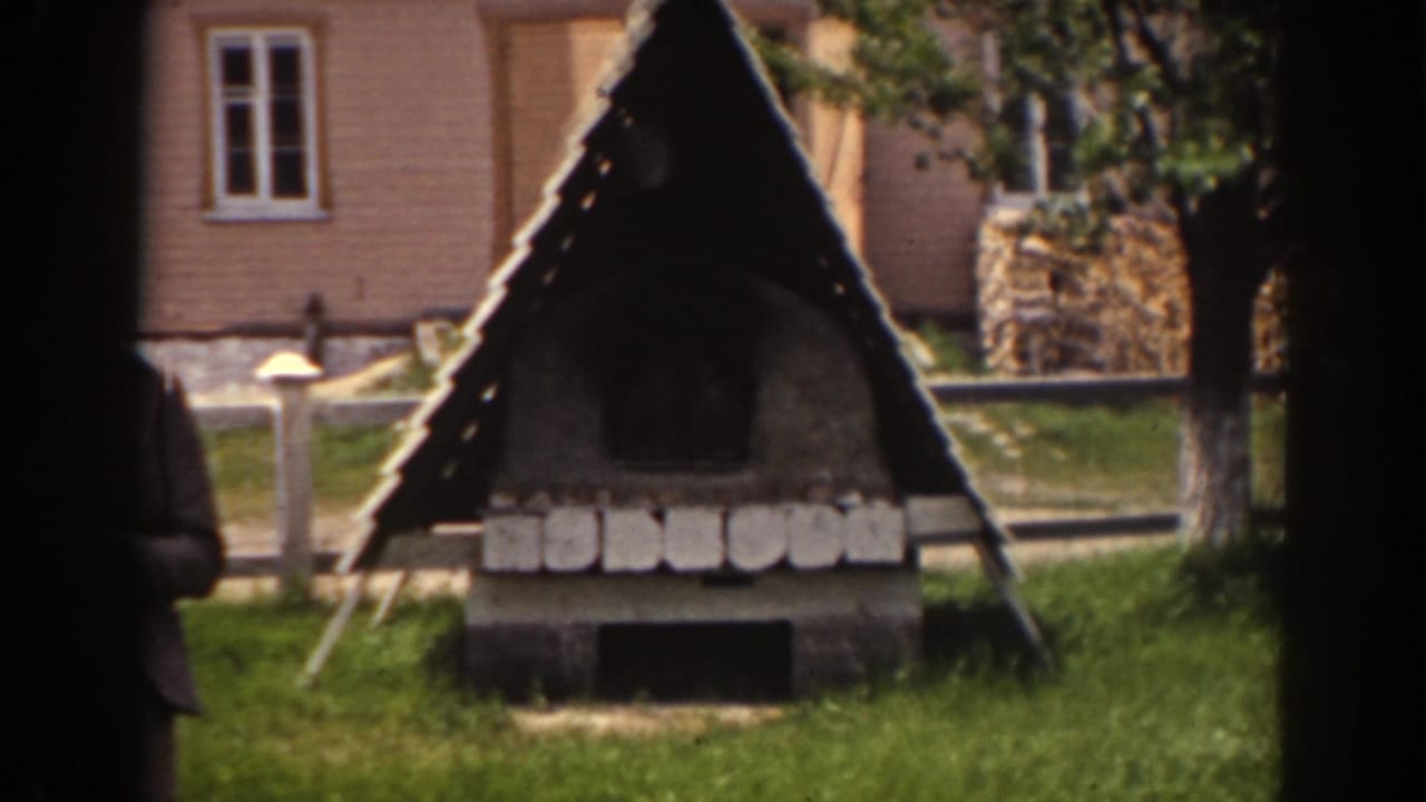1938 Mount Washington frame with a roofed stone structure in grass, backed by a fence, pale house wall, windows, and trees.