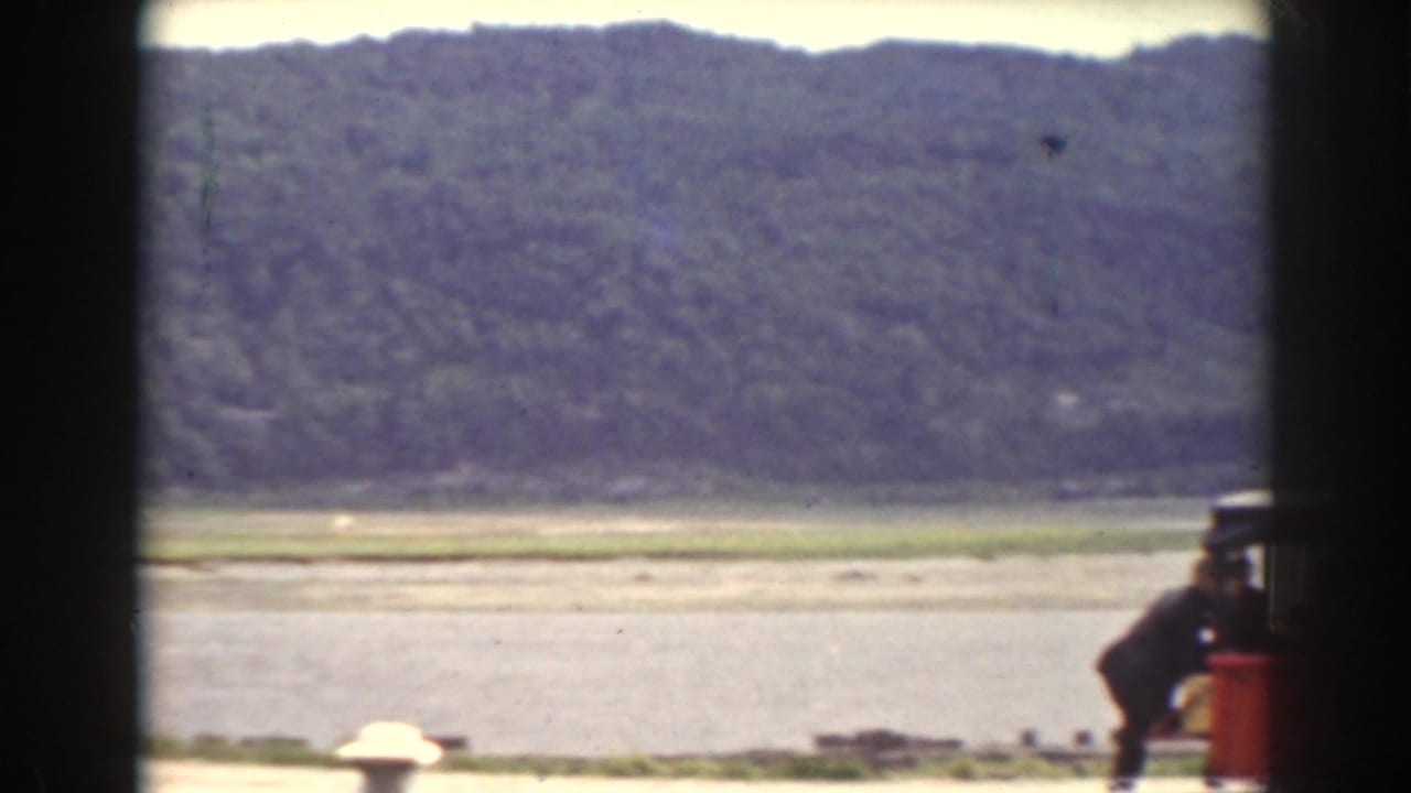 1938 Mount Washington-area frame looking across water toward wooded hills, with part of a red vehicle entering the foreground.
