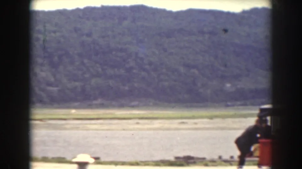 1938 Mount Washington-area frame looking across water toward wooded hills, with part of a red vehicle entering the foreground.