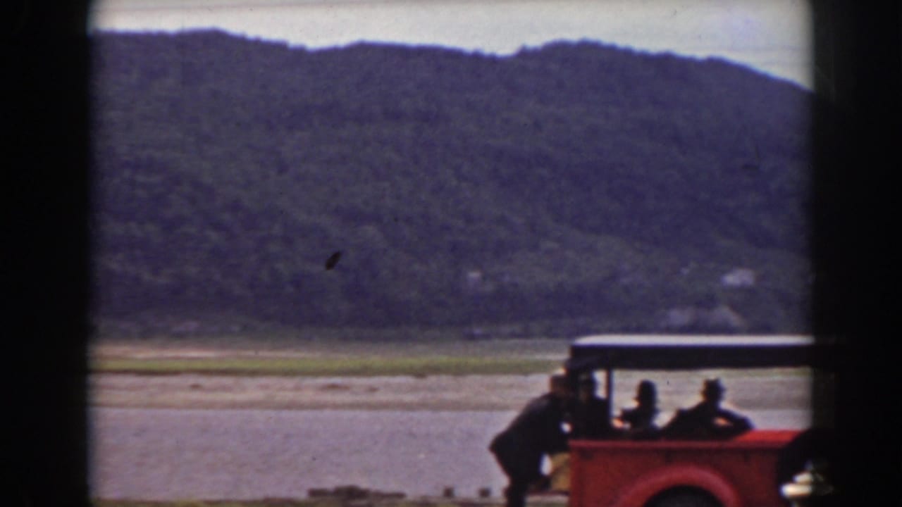 1938 Mount Washington-area home movie frame showing people beside a red open vehicle near water, with wooded hills behind them.
