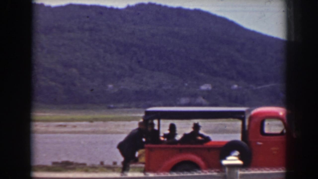 Red pickup truck with passengers beside a lake and wooded mountain.
