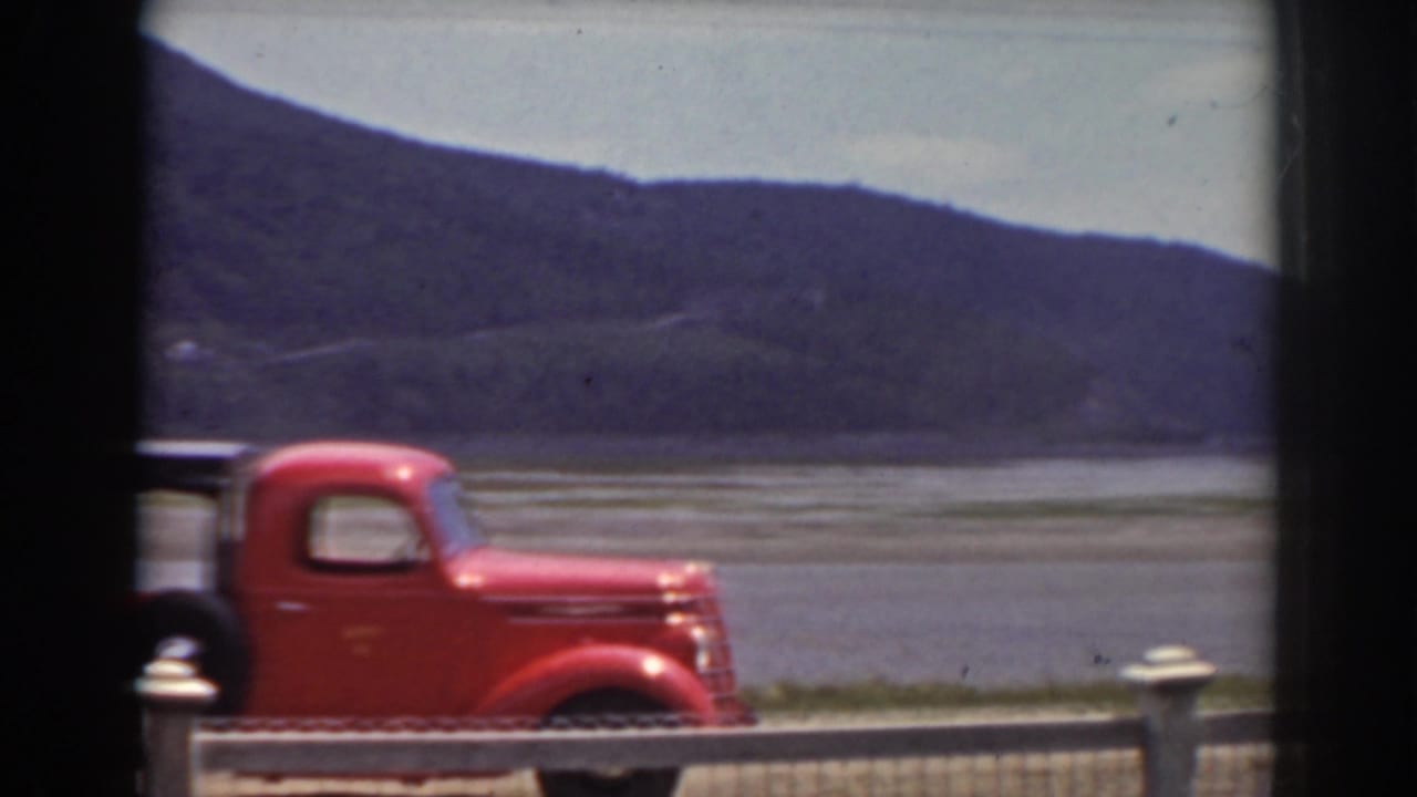 The red pickup exits the frame along a lakeside road, with Mount Washington-area hills and water visible behind it.
